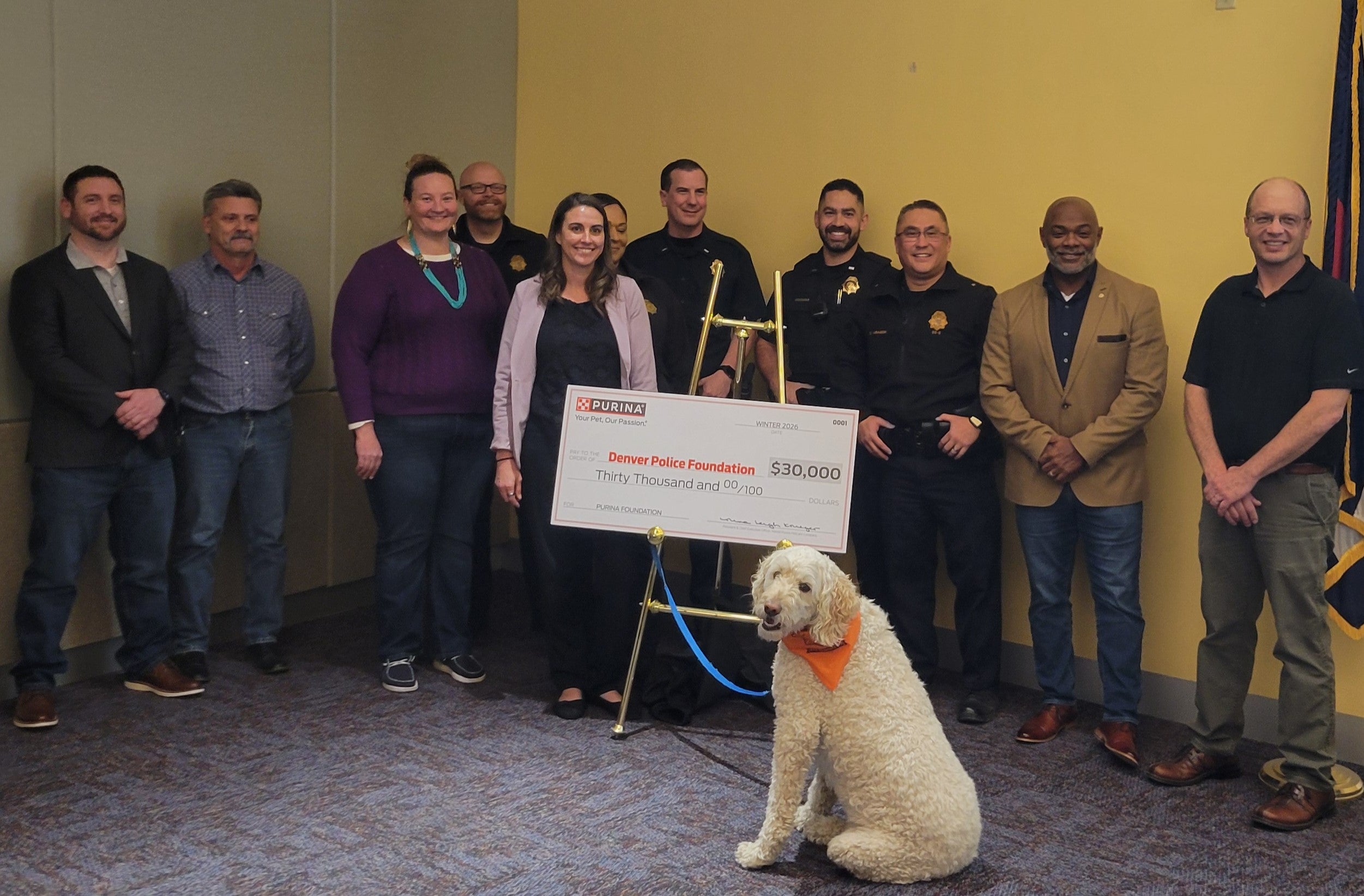 Group of people standing behind a dog wearing an orange bandana and a $30,000 donation check for the Denver Police Foundation