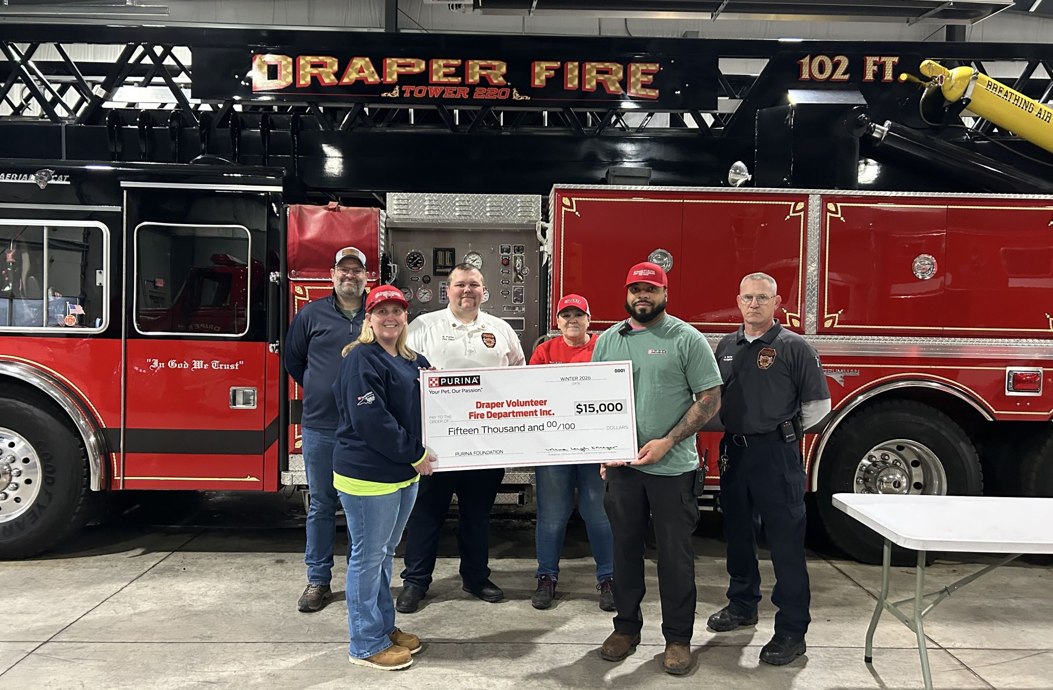 Group of six people holding a $15,000 donation check in front of a red Draper Fire Department fire truck inside the station.
