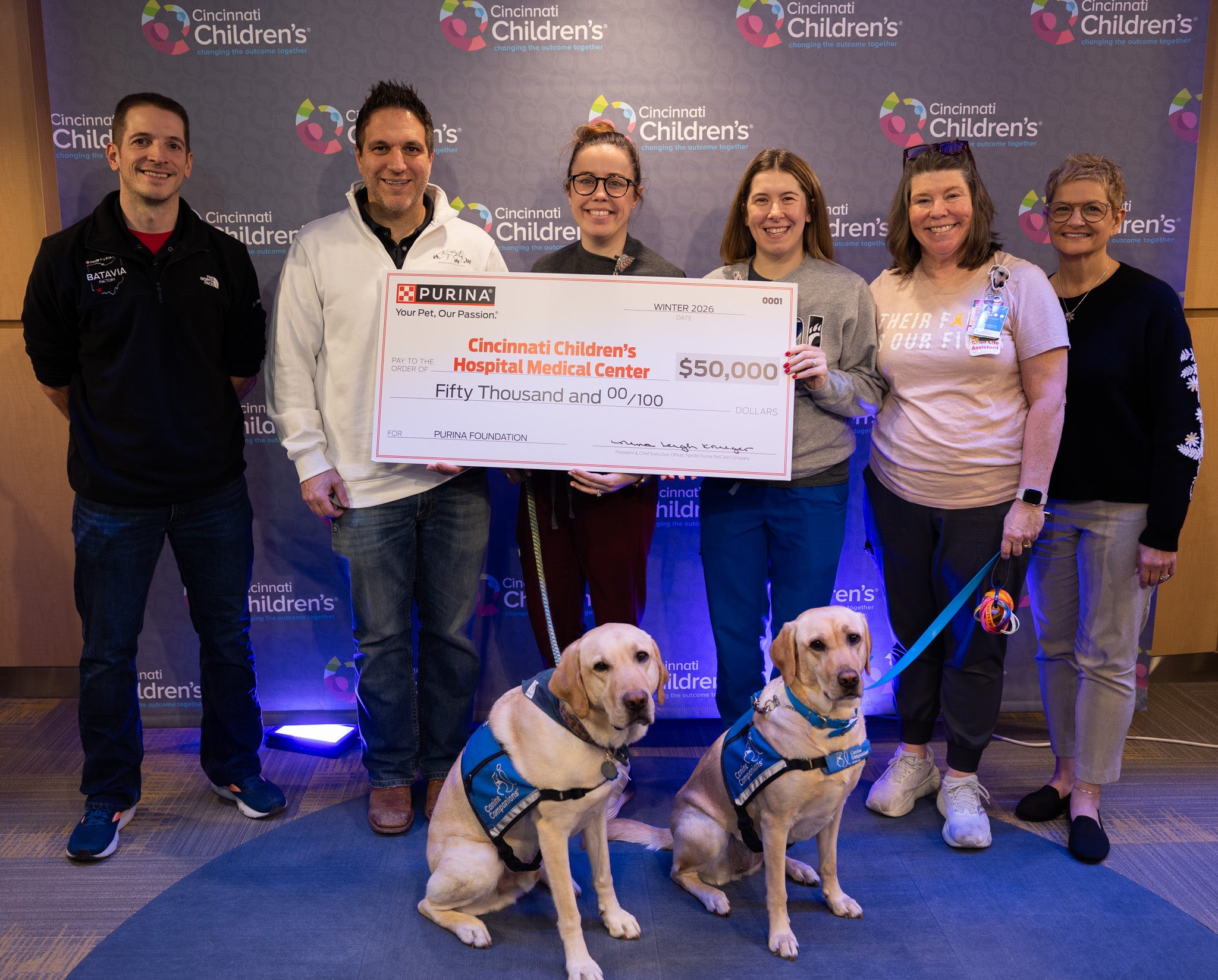 Group of six people holding a $50,000 donation check to Cincinnati Children's Hospital, accompanied by two service dogs weari