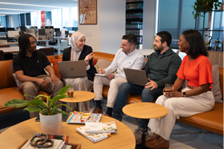 Five coworkers sit on a couch in an office, smiling and talking with laptops and notebooks.
