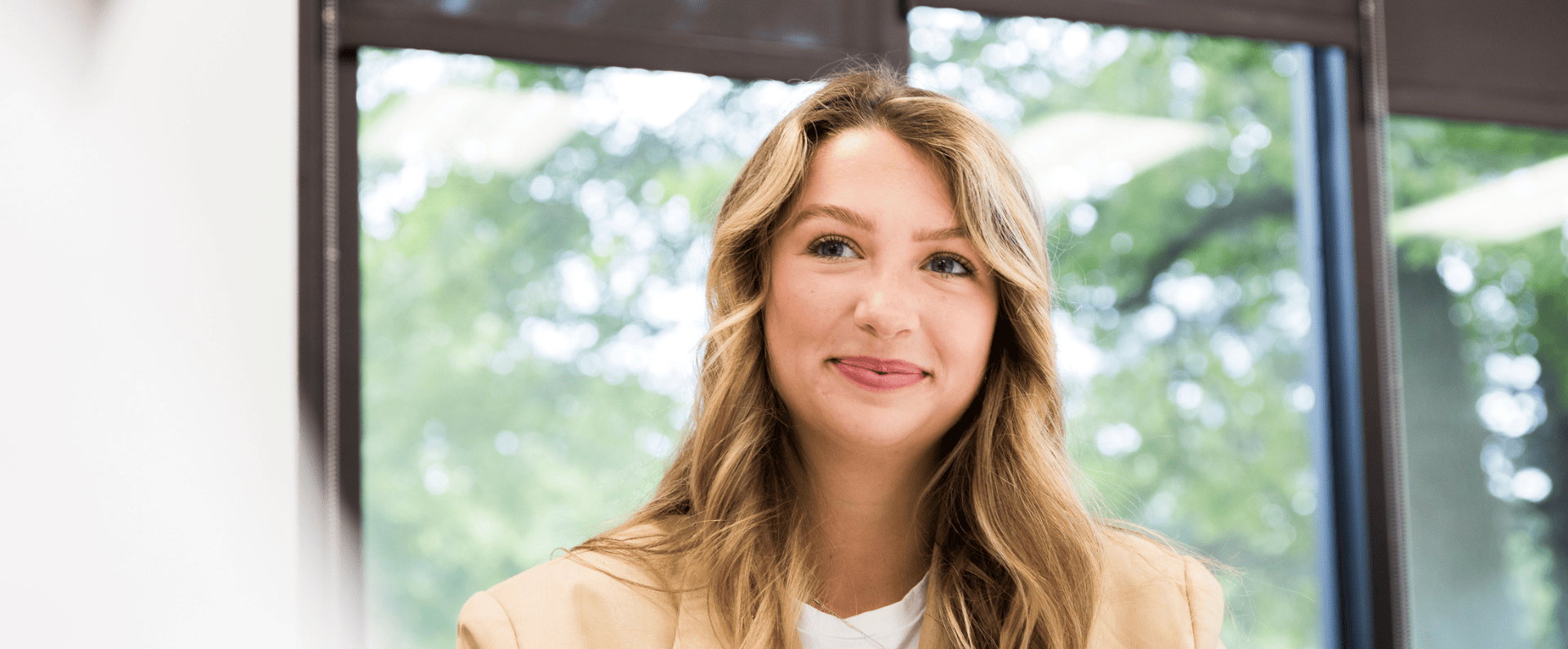 Young woman with long blonde hair wearing a beige blazer and white shirt, sitting indoors with large windows and greenery out