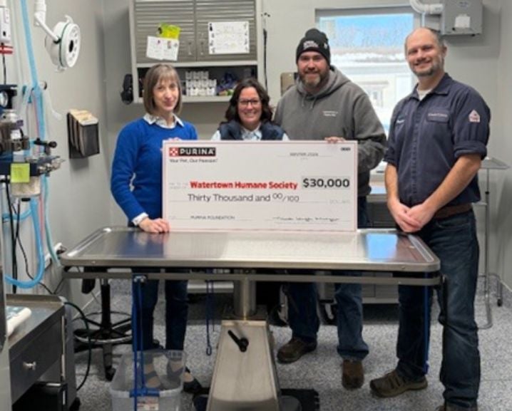 Four people stand behind a veterinary exam table holding a $30,000 donation check to Watertown Humane Society from Purina Fou