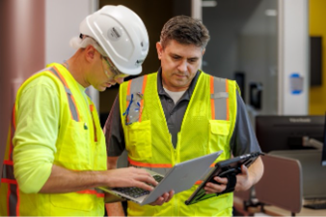 Two construction workers in safety vests reviewing information on a laptop and tablet.