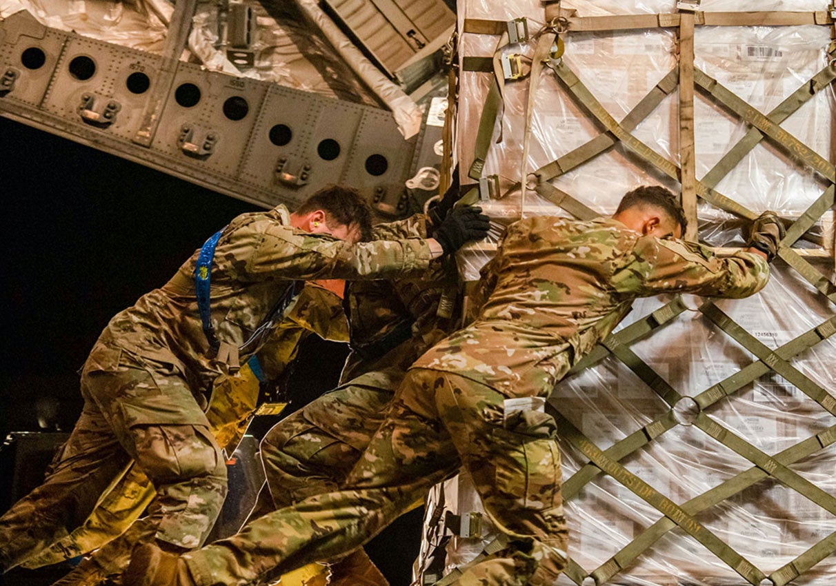 Three soldiers in camouflage uniforms push a large cargo pallet inside an aircraft.