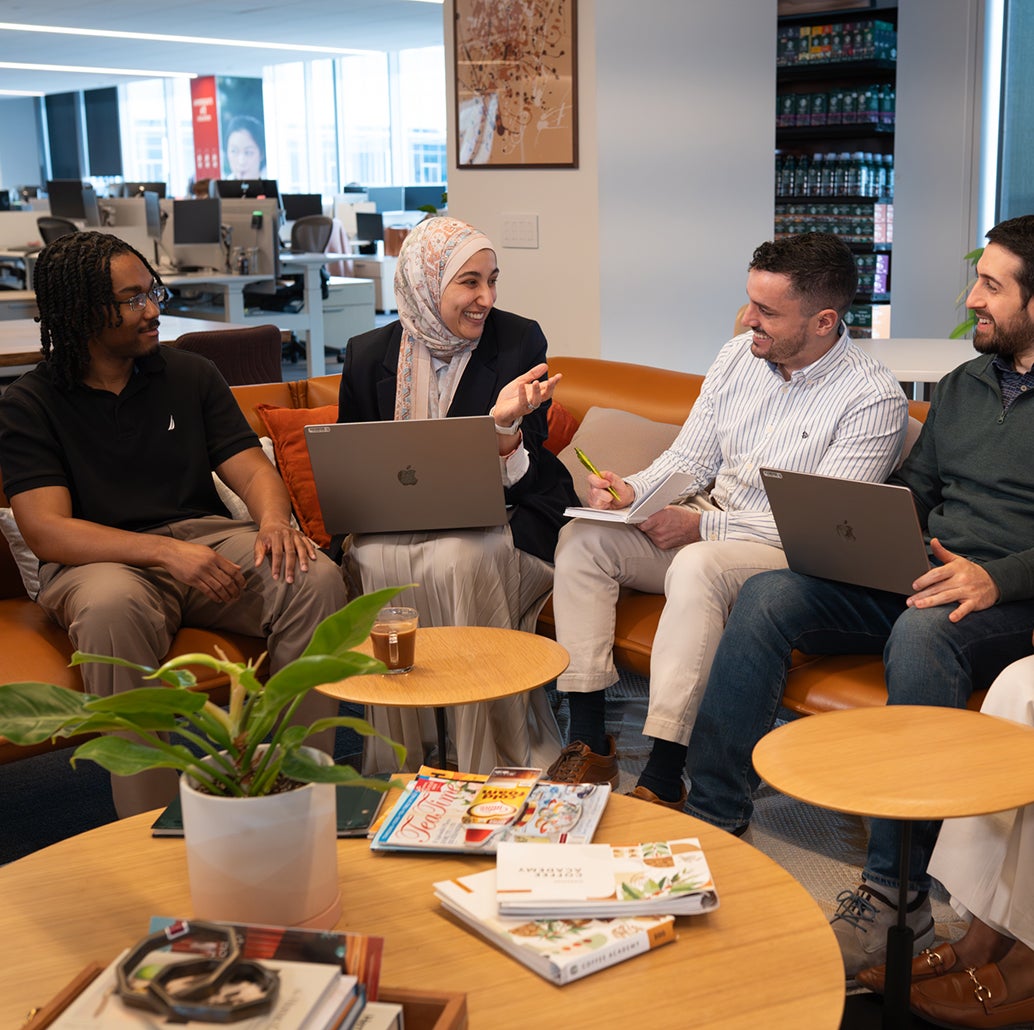 Five colleagues sit on a couch in an office, talking and smiling with laptops and notes.