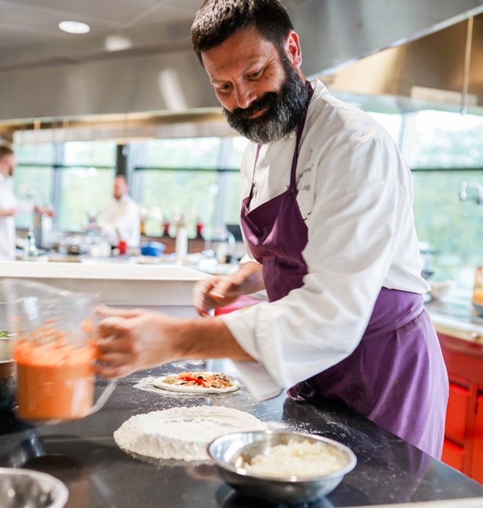 Chef in a purple apron preparing pizza dough with sauce in a modern kitchen.
