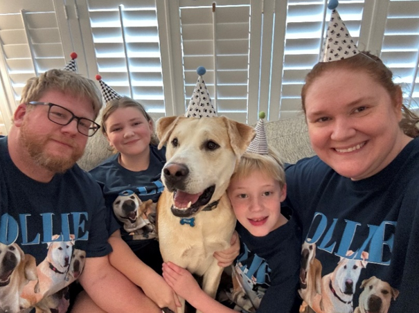 Family wearing matching dog-themed shirts and party hats, celebrating with a happy yellow Labrador in the center.