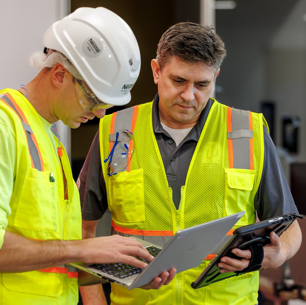 Two construction workers in safety vests look at a laptop and tablet.