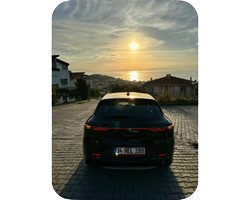Black car parked on a cobblestone street facing a sunset over the ocean, with houses and hills in the background.