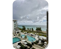 View of a hotel pool area with lounge chairs, palm trees, and the ocean under a cloudy sky.