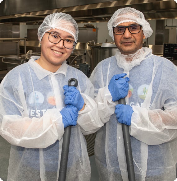 Two people in hairnets and gloves standing in a commercial kitchen, smiling.