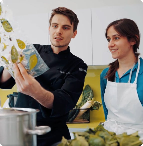 Chef showing herbs in a plastic bag to a smiling woman in a kitchen.