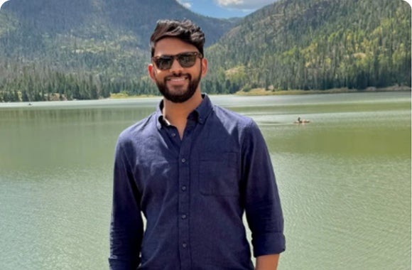 Man in sunglasses smiling by a lake with forested hills in the background.