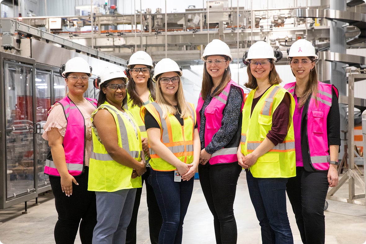 Seven women in safety vests and hard hats smile in a factory setting.