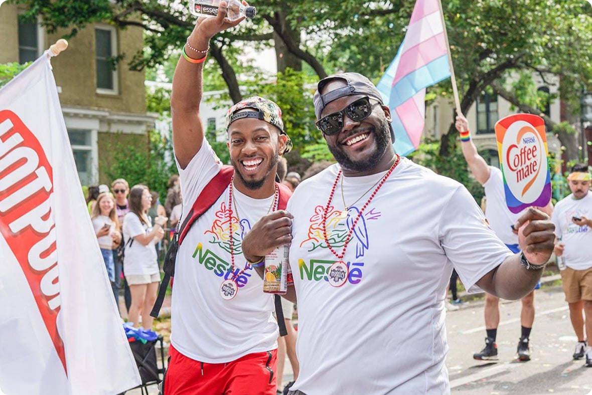 Two smiling men at a parade wearing rainbow Nestlé shirts, surrounded by festive crowd and flags.