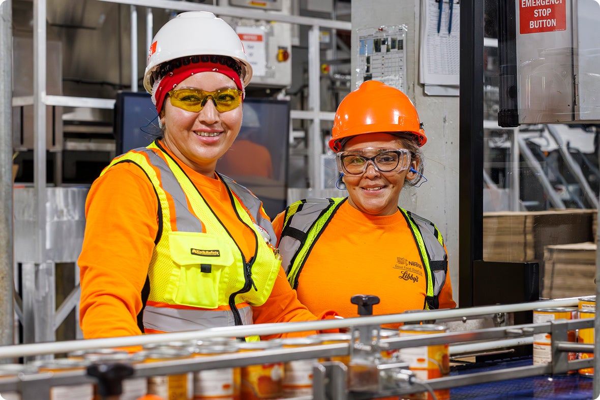 Two women in safety gear and hard hats smiling in a factory setting.