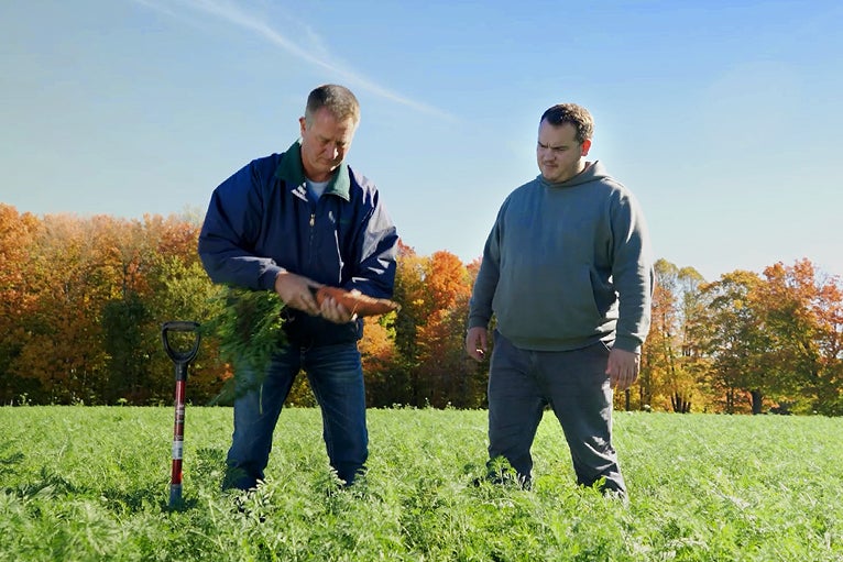 Two men in a field, one holding freshly harvested carrots, trees with autumn foliage behind.