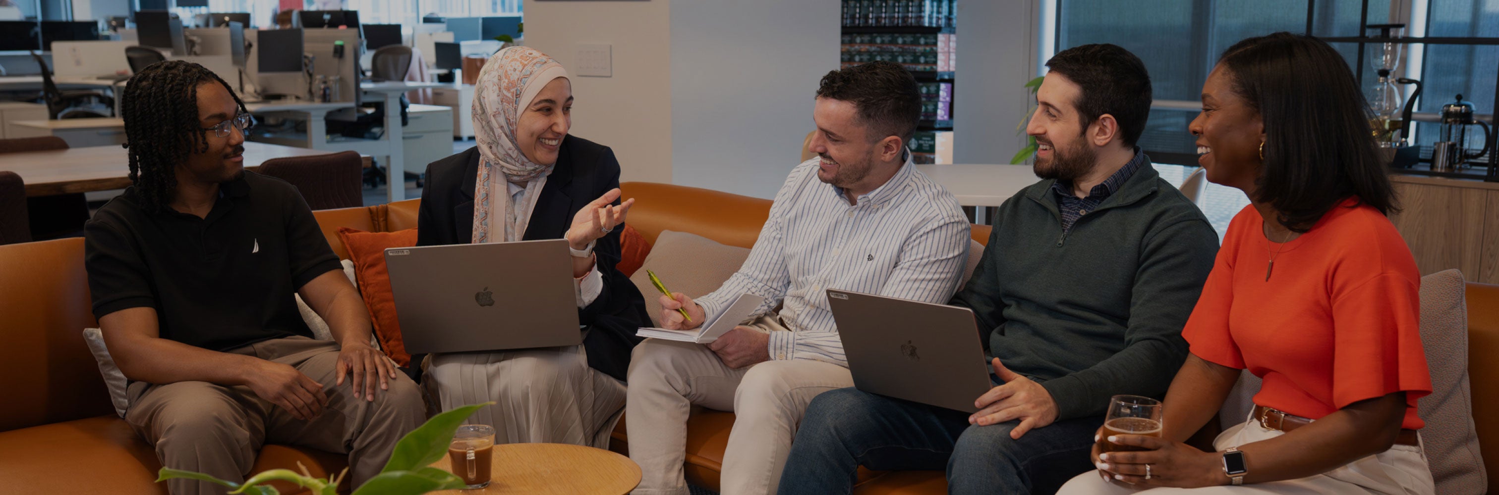 Five colleagues sit together on a couch, smiling and talking in an office setting.