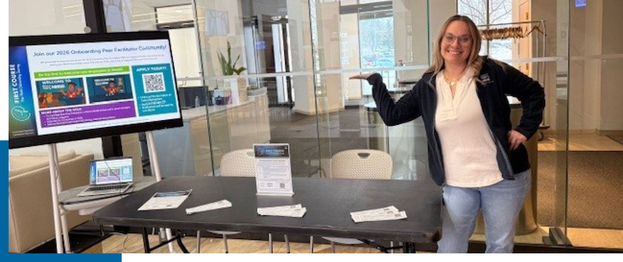 Smiling woman stands by an information table with brochures and a display screen.