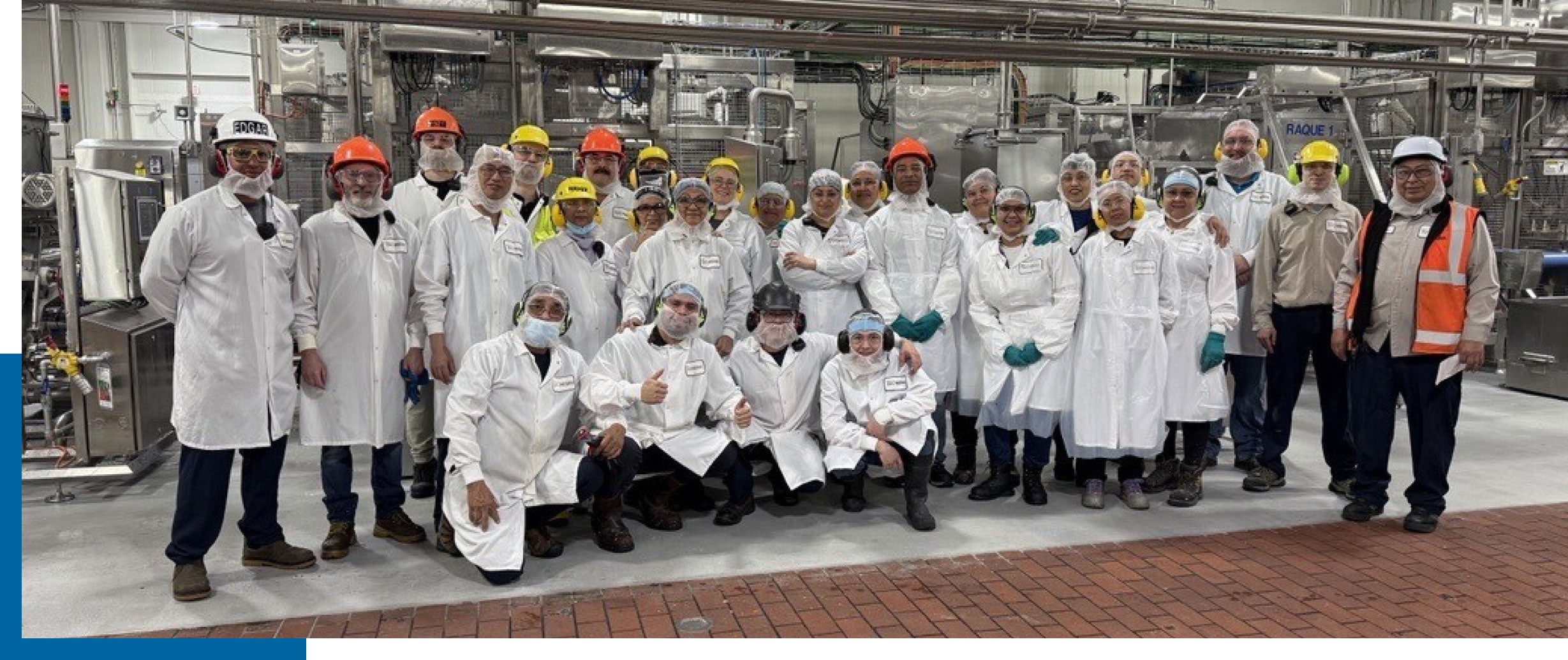 Group of factory workers in lab coats and helmets posing in an industrial facility.