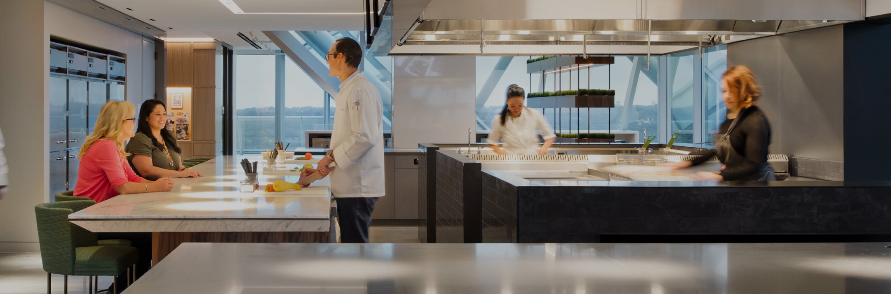 Modern kitchen with chefs preparing food and two women seated at a counter.