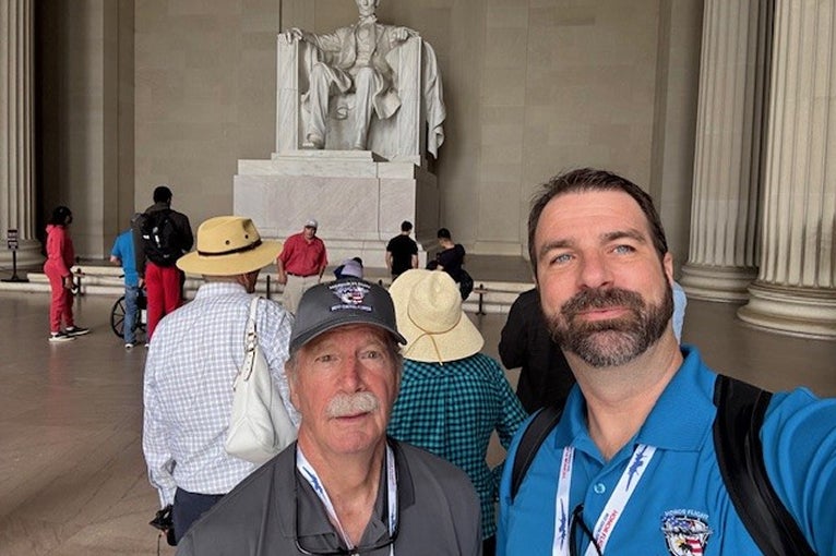 Two men pose for a selfie at the Lincoln Memorial, surrounded by visitors.