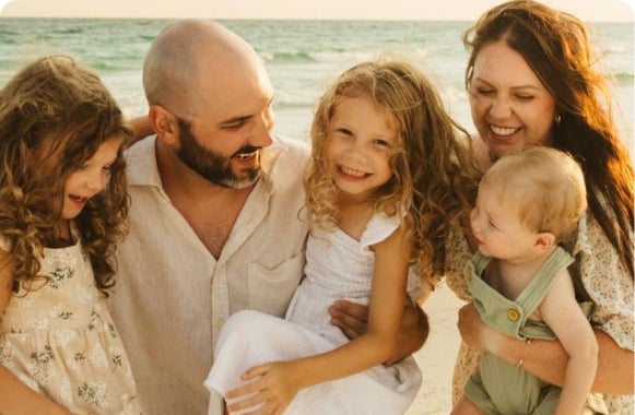 Family smiling together on a sunny beach, with ocean waves in the background.