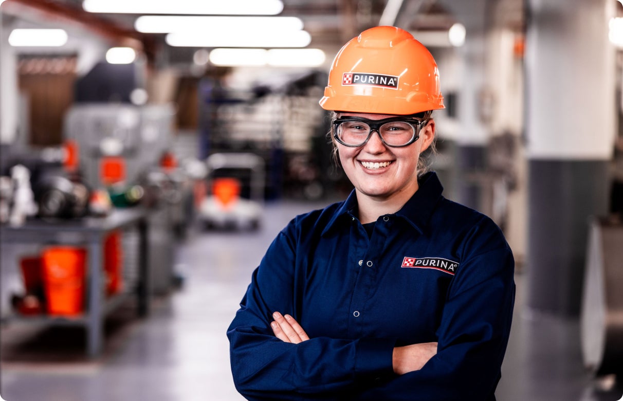 Smiling worker in safety gear and Purina uniform in an industrial setting.