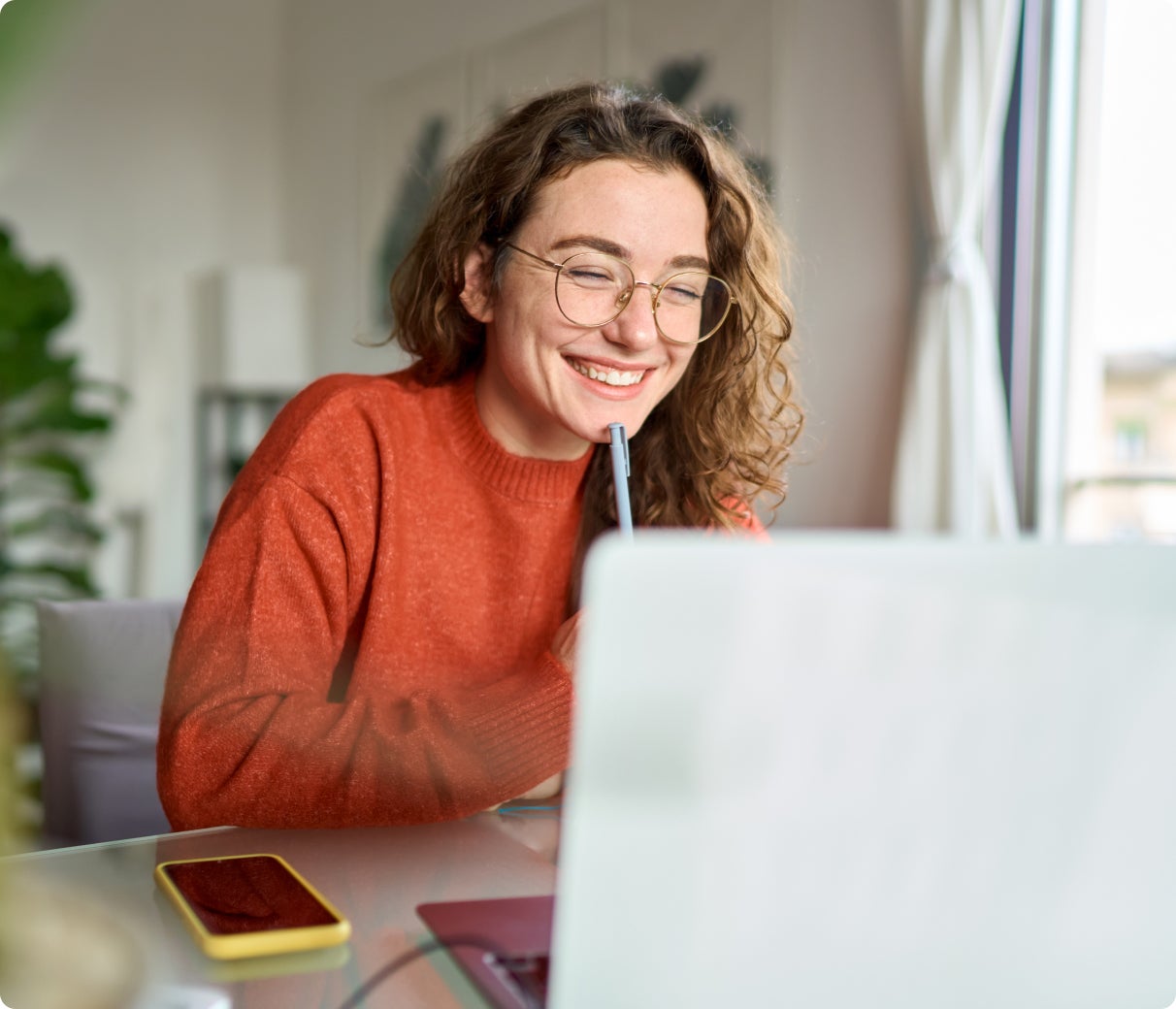Woman with glasses smiling at laptop, holding pen, seated by a window.