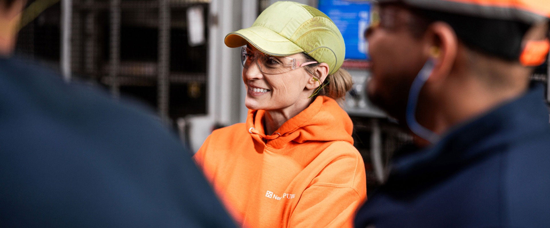 Smiling woman in orange hoodie and safety gear talking with coworkers in an industrial setting.