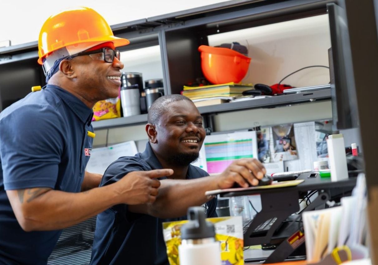 Two construction workers collaborate at a workstation, with safety helmets, tools, and equipment visible behind them.