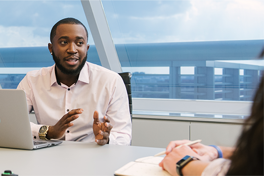 Employee sitting at a desk with his laptop talking with a colleague