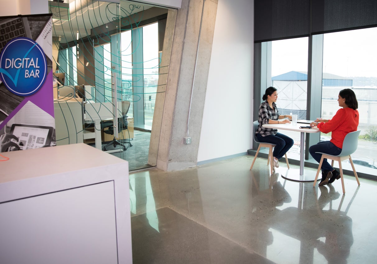 Two employees sitting next to window next to the digital bar inside an office setting 