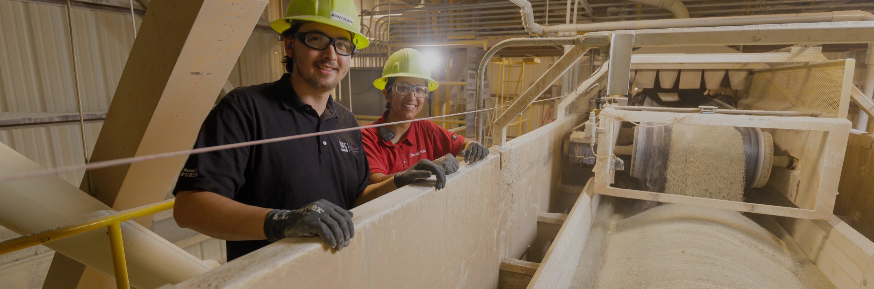 Two men in hard hats stand beside a conveyor belt, engaged in conversation about the ongoing work process.