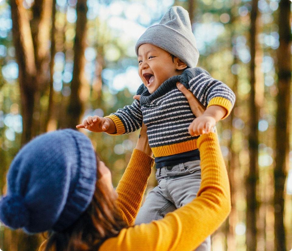 Adult lifting a laughing baby in a striped sweater and hat outdoors among trees.