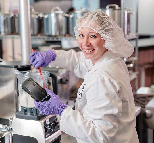 Smiling lab worker with gloves and hairnet mixing ingredients in a blender