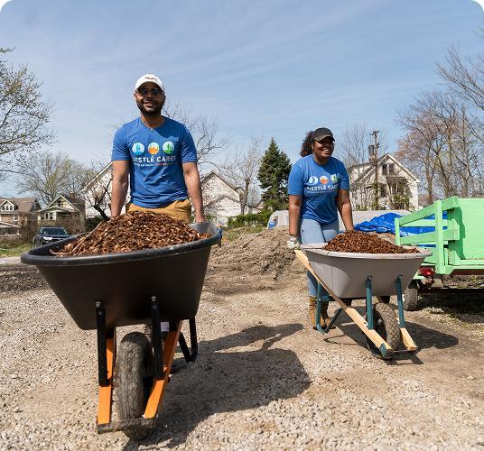 Two people with wheelbarrows of mulch working outdoors on a sunny day.