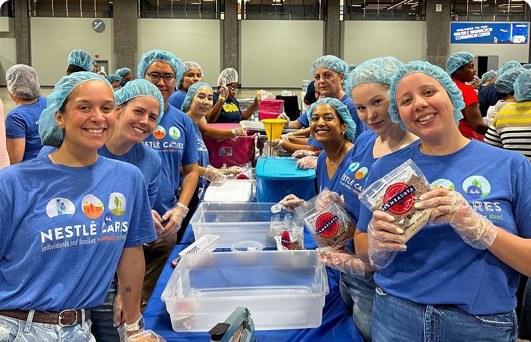 Group of smiling volunteers in blue shirts and hairnets packing food at a community event.