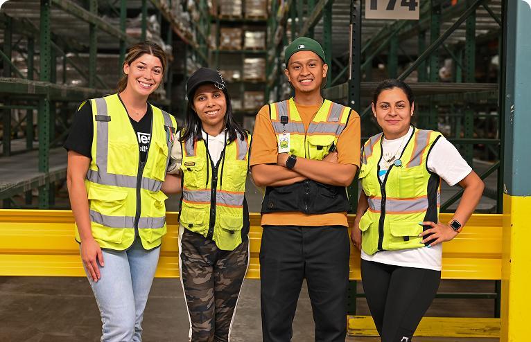 Four warehouse workers in reflective vests smiling and posing together in a warehouse aisle.