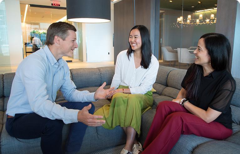 Three people sitting on a couch, smiling and having a casual conversation in an office.