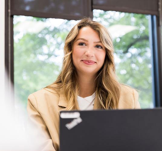 Smiling woman with long hair sits at a desk, laptop open, window and trees behind her.