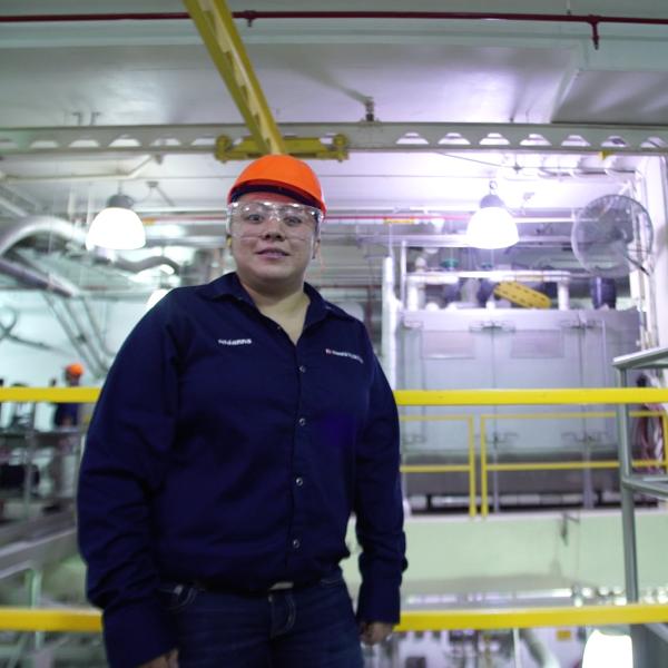 Woman in factory with blue jacket, clear safety goggles and orange safety hat