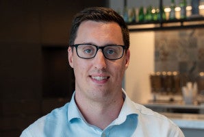 Man with short brown hair and glasses smiling indoors, bottles on shelves behind him.