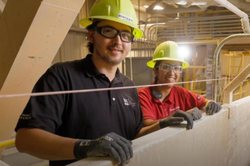 Two individuals in hard hats and safety glasses stand beside a wall, ready for a construction project.