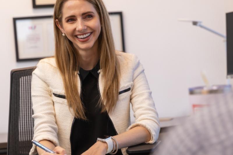 A woman seated at a desk, holding a pen, appears focused and engaged in her work.