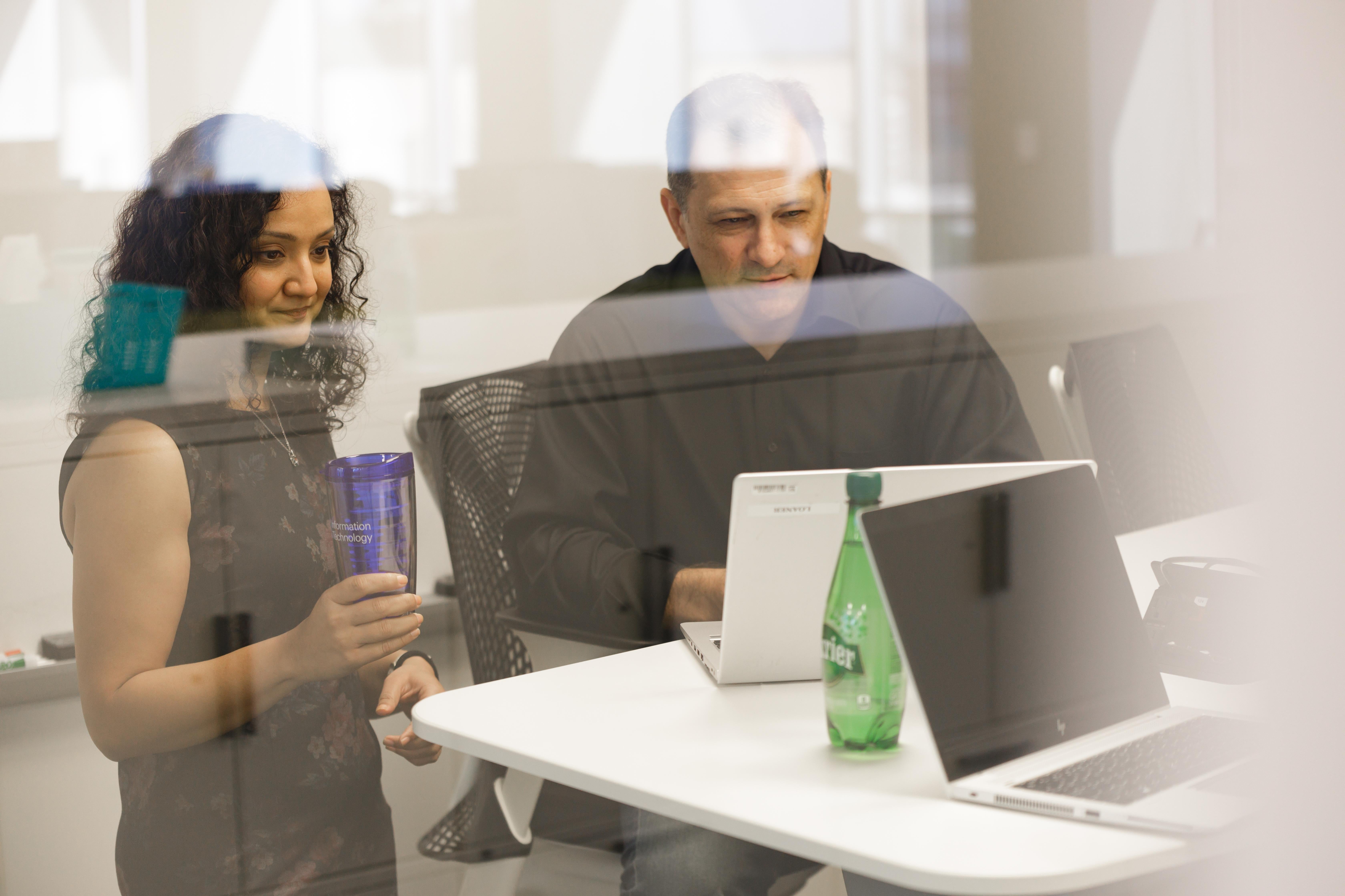 Woman standing and man seated at a desk with laptops and Perrier bottle.