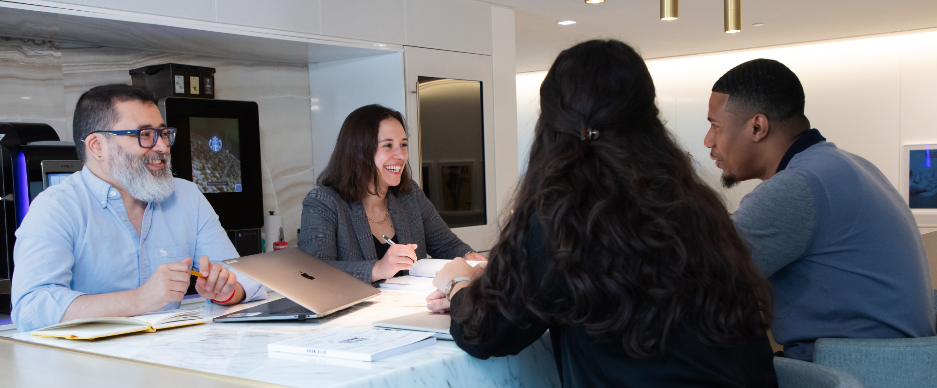 Four professionals in a modern office kitchen having a meeting around a marble table with laptops and notebooks.
