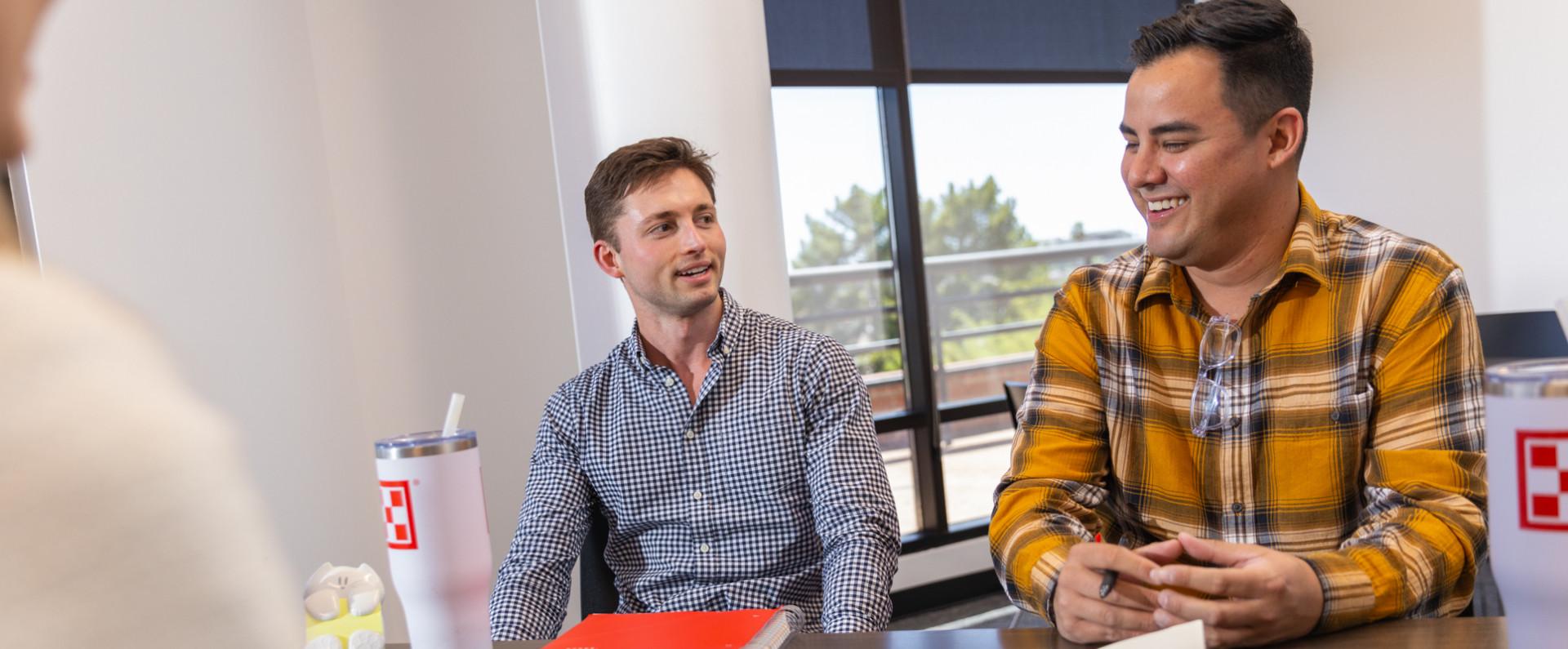 Two men sit at a table in a bright office, engaged in conversation during a meeting, with drinks and notebooks